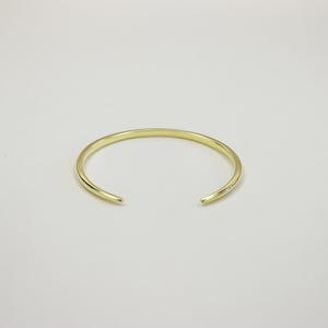 May include: A polished gold-tone cuff bracelet with a smooth, rounded surface and a simple open-ended design. The metal band features a consistent thickness throughout its circular shape, creating a clean and reflective appearance for the wrist.