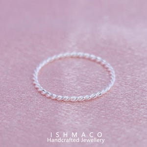 Op de afbeelding: Een zilverkleurige, gedraaide touwring. De ring staat tegen een zachtroze achtergrond. De tekst "ISHMACO Handcrafted Jewellery" staat onderaan de afbeelding.