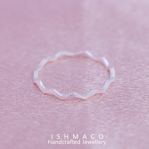 Puede incluir: Un anillo de plata con un diseño ondulado, sobre un fondo rosa suave. La superficie del anillo refleja la luz, resaltando su patrón en zigzag. El anillo es una pieza de joyería artesanal.