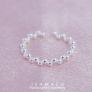 Puede incluir: Un anillo plateado con un diseño de cuentas. El anillo está abierto en la parte superior y presenta pequeñas cuentas redondas que crean un patrón texturizado y ondulado. El anillo está sobre un fondo rosa suave. El texto "ISHMACO Handcrafted Jewellery" está en la parte inferior.