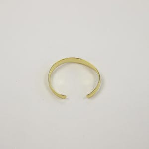 May include: A polished gold-tone cuff bracelet with a smooth, reflective surface and a simple open-ended design. The bracelet features a rounded band that curves into a circular shape, maintaining a consistent width throughout its entire length.
