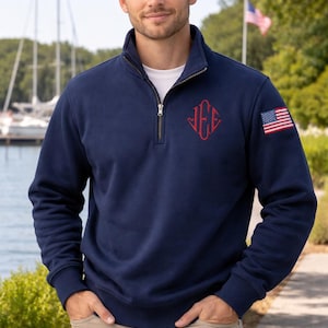Könnte beinhalten: Marineblaues Sweatshirt mit Stehkragen, rotem Monogramm und einem amerikanischen Flaggenaufnäher am Ärmel. Das Sweatshirt wird über einem weißen T-Shirt getragen. Der Hintergrund umfasst ein Gewässer und Segelboote.