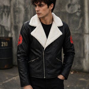May include: Black faux leather jacket with a white faux fur collar. The jacket has a zip closure, two side pockets, and red flame designs on the sleeves. The model is wearing a black shirt and black trousers.