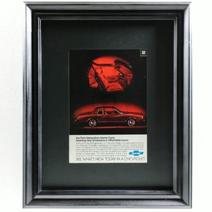 May include: Framed vintage advertisement for a Chevrolet Monte Carlo. The ad features a red car and interior, with text that reads "SEE WHAT'S NEW TODAY IN A CHEVROLET". The frame is black.