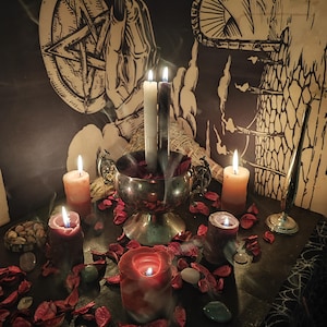 May include: A mystical scene featuring lit candles of various sizes and colors, including white, orange, and red. A silver bowl holds burning candles and red petals. The background includes a pentagram and other occult symbols, creating an atmospheric setting.