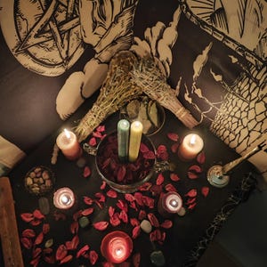 May include: A still-life arrangement featuring lit candles, dried flowers, and sage bundles. The scene includes a glass bowl with red petals, two green and yellow candles, and scattered stones. A black and white tapestry is in the background.