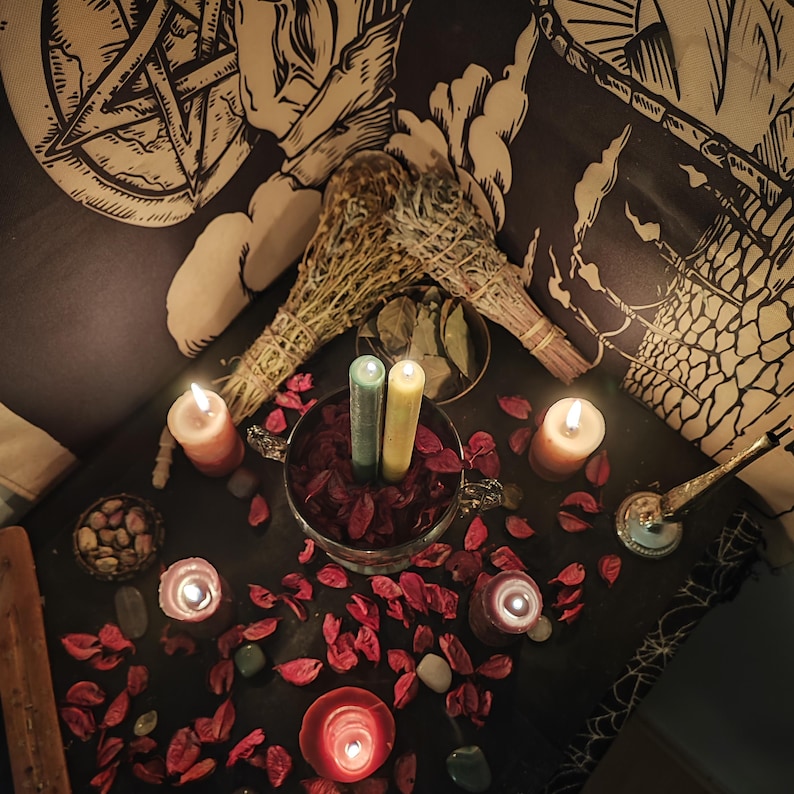 May include: A still-life arrangement featuring lit candles, dried flowers, and sage bundles. The scene includes a glass bowl with red petals, two green and yellow candles, and scattered stones. A black and white tapestry is in the background.