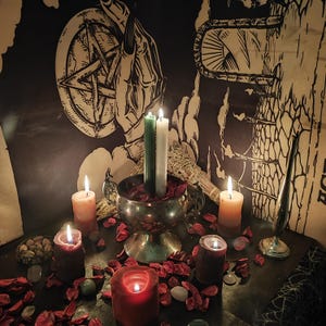 May include: A mystical scene featuring lit candles of various colors, including red, orange, and green, arranged on a dark surface. A silver chalice holds candles and dried petals. A pentagram and other occult symbols are visible in the background.