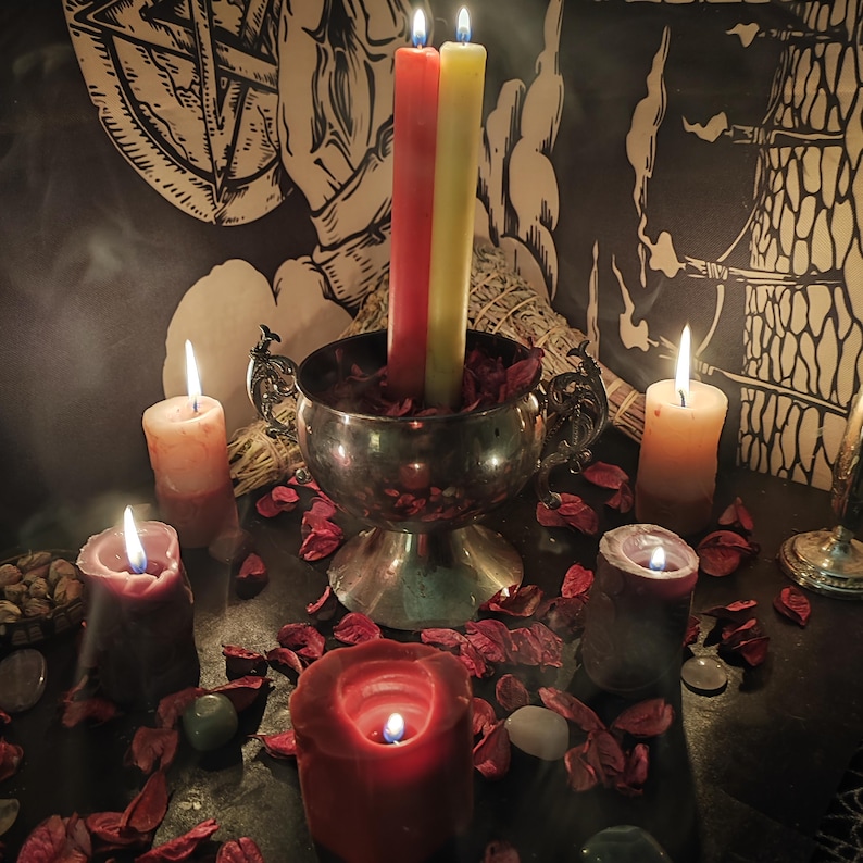 May include: A mystical scene featuring lit candles of various sizes and colors, including red, yellow, and orange. A silver bowl holds two tall candles, surrounded by dried rose petals and other ritualistic items. A pentagram design is visible in the background.