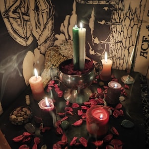 May include: A dimly lit scene featuring lit candles of various sizes and colors, including green, white, orange, and red. A silver bowl holds two candles and red petals. The scene evokes a mystical or spiritual theme.