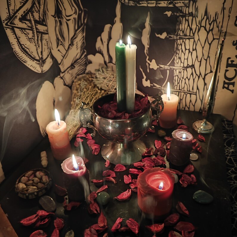 May include: A dimly lit scene featuring lit candles of various colors and sizes. A silver chalice holds two candles, one green and one white, surrounded by red petals. Other candles are orange, red, and purple. The scene evokes a mystical or spiritual theme.