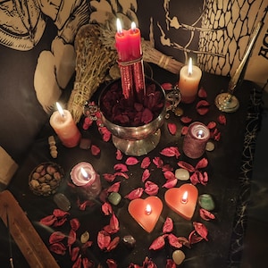 May include: A dimly lit scene featuring lit candles of various shapes and sizes, including heart-shaped and cylindrical ones. A silver bowl filled with red petals holds two red candles. Dried herbs and scattered crystals add to the mystical ambiance.