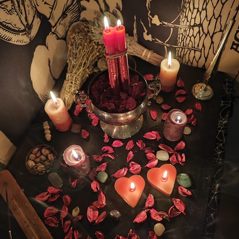 May include: A dimly lit scene featuring lit candles of various shapes and sizes, including heart-shaped and cylindrical ones. A silver bowl filled with red petals holds two red candles. Dried herbs and scattered crystals add to the mystical ambiance.