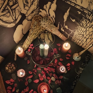 May include: A dimly lit table with candles, dried herbs, and decorative items. Two bundles of dried herbs are placed near a metal bowl with red petals and two lit candles. Several other candles and small stones are scattered around the table.