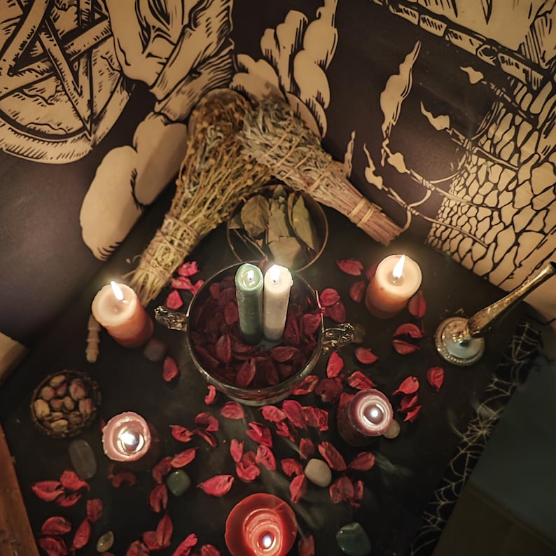 May include: A dimly lit table with candles, dried herbs, and decorative items. Two bundles of dried herbs are placed near a metal bowl with red petals and two lit candles. Several other candles and small stones are scattered around the table.