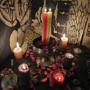 May include: A collection of lit candles in various sizes and colors, including red, yellow, and orange. The candles are arranged on a dark surface with scattered red petals and small green stones. A silver chalice holds two tall candles.
