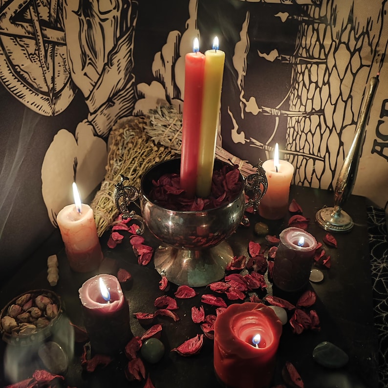 May include: A collection of lit candles in various sizes and colors, including red, yellow, and orange. The candles are arranged on a dark surface with scattered red petals and small green stones. A silver chalice holds two tall candles.
