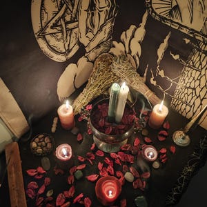 May include: A mystical scene featuring lit candles of various sizes and colors, including orange, red, and green. A silver bowl holds red petals and two candles. Dried sage bundles and a pentagram backdrop add to the spiritual ambiance.
