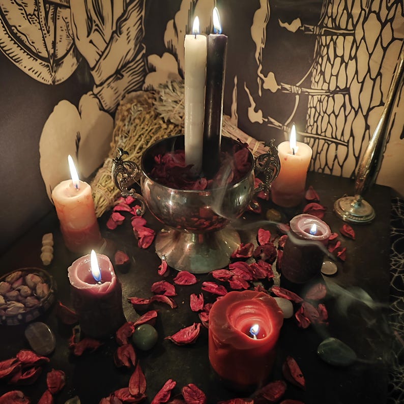 May include: A mystical scene featuring lit candles of various sizes and colors, including white, burgundy, and orange. A silver chalice holds two candles, surrounded by red petals and green stones. Smoke rises, creating an atmospheric ambiance.