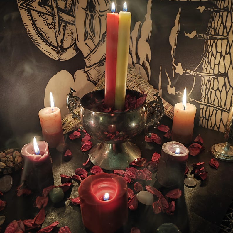 May include: A mystical scene featuring lit candles of various sizes and colors, including red, yellow, and orange. A silver bowl holds two tall candles, surrounded by dried rose petals and other ritualistic items. A pentagram design is visible in the background.