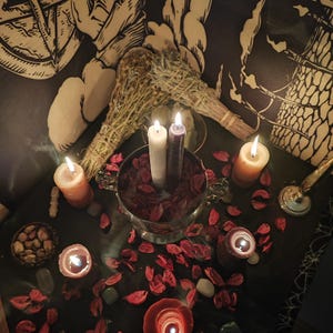 May include: A mystical scene featuring lit candles, dried herbs, and a silver bowl filled with red petals. The candles vary in color, with a white and purple candle in the center. The scene is set on a dark surface, with a patterned backdrop.