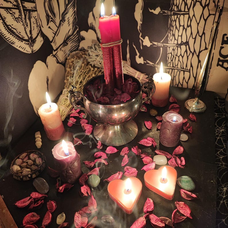 May include: A mystical arrangement featuring lit candles of various shapes and sizes, including heart-shaped and cylindrical ones. A silver chalice holds red rose petals and two tall red candles. The scene is adorned with dried herbs and scattered petals, creating a spiritual ambiance.