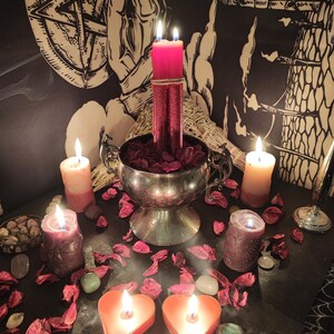 May include: A mystical arrangement featuring lit candles of various sizes and colors, including heart-shaped and cylindrical candles. A silver bowl holds red petals, with additional petals scattered around. The scene is set against a backdrop of esoteric artwork and dried herbs.