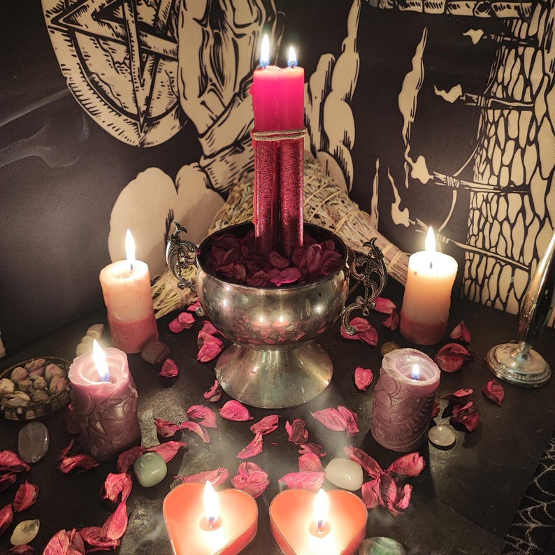 May include: A mystical arrangement featuring lit candles of various sizes and colors, including heart-shaped and cylindrical candles. A silver bowl holds red petals, with additional petals scattered around. The scene is set against a backdrop of esoteric artwork and dried herbs.