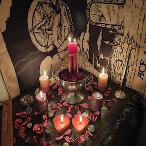 May include: A mystical altar setup featuring lit candles of various sizes and colors, including heart-shaped ones. A silver bowl holds red rose petals, and a pentagram tapestry hangs in the background. The scene is illuminated with warm light.