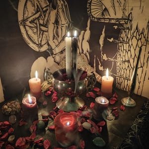 May include: A mystical scene featuring lit candles of various sizes and colors, including red, orange, and white. A silver bowl holds burning candles and is surrounded by red rose petals. The background includes a pentagram and other occult symbols.