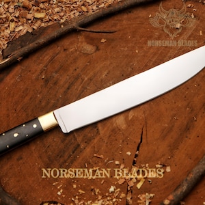 May include: A large, stainless steel chef's knife with a black handle adorned with gold-colored accents. The blade is curved and sharp, and the knife is set against a wooden surface. The text "NORSEMAN BLADES" is visible.
