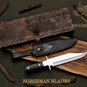 May include: A hunting knife with a polished steel blade and black handle, resting on a wooden cutting board. The knife is accompanied by a black leather sheath and a wooden box with a wolf engraving. The words "NORSEMAN BLADES" are visible.