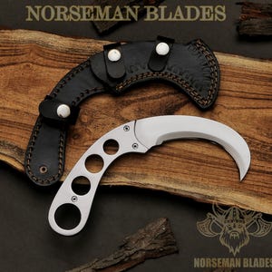 Op de afbeelding: Een gebogen, zilverkleurig karambitmes met een handvat met drie cirkelvormige uitsparingen, samen met een zwarte leren schede. Het mes en de schede worden gepresenteerd op een houten oppervlak. De tekst "NORSEMAN BLADES" is zichtbaar.