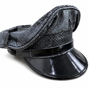 Peut inclure: Une casquette de style militaire à motif peau de serpent gris, dotée d'une calotte haute et d'une visière structurée en cuir verni noir brillant. Une fine bride noire brillante traverse la base de la calotte, fixée par des boutons noirs.