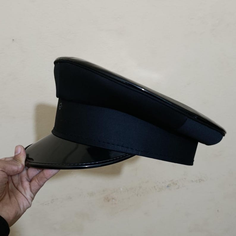 May include: A black, glossy visor hat with a structured crown and a dark band. The hat has a shiny, patent leather-like brim and a button detail on the side. The hat is held by a hand.