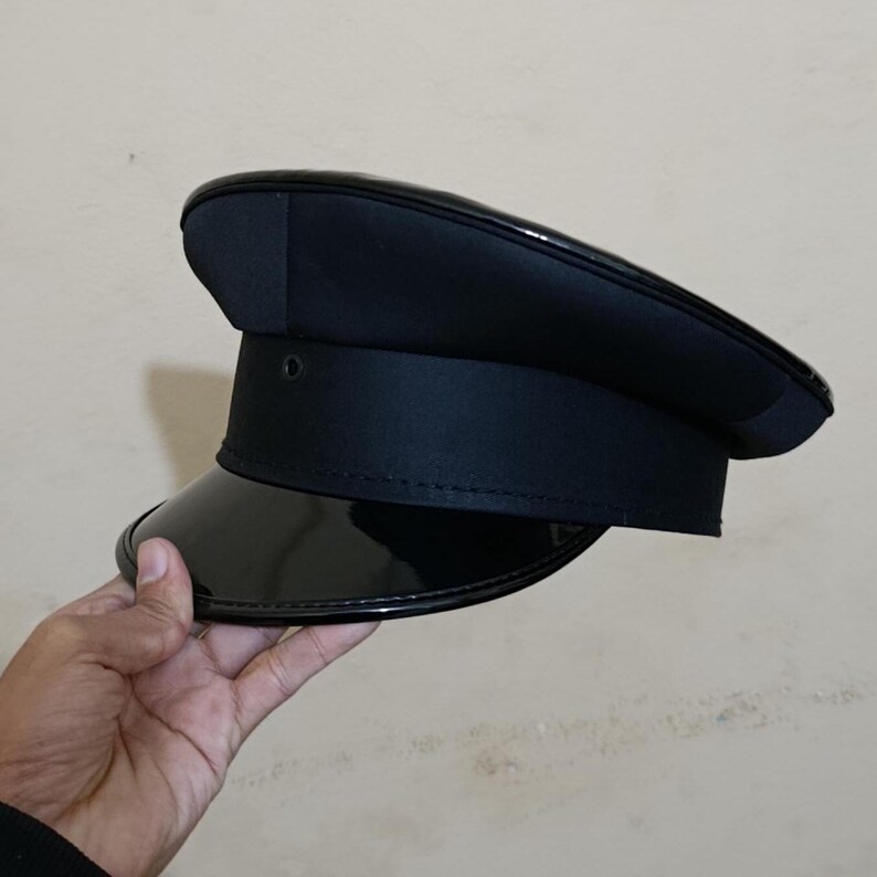 May include: A black hat with a shiny, patent leather visor and a structured crown. The hat has a band around the base and a small, circular detail on the side. The hat is held by a hand.