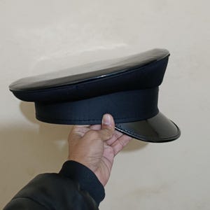 May include: A black, glossy, military-style hat with a patent leather visor and a structured crown. The hat has a band around the base and is held by a hand. The hat is a classic accessory.