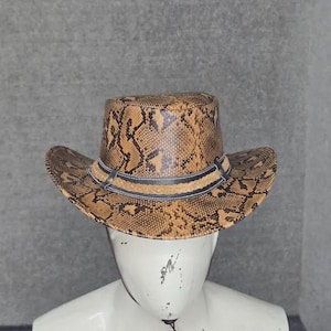 Op de afbeelding: Een cowboyhoed met een beige en bruin slangenleerpatroon over de kroon en de rand. De hoed is voorzien van een donkerbruine band met een gevlochten detail in het midden die de basis van de kroon omsluit voor een gestructureerde uitstraling.