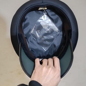 May include: Black hat with a rounded crown and a stiff, dark green visor. The interior is lined with black fabric and features a clear, diamond-shaped tag. The hat is being held up, revealing its inner construction.