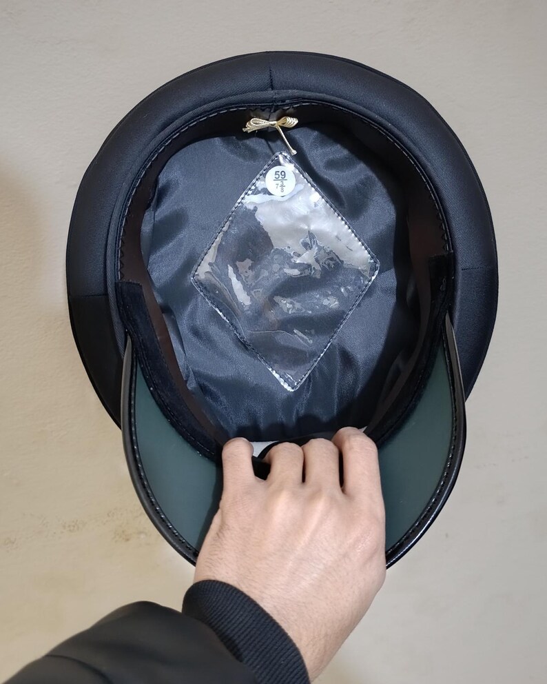 May include: Black hat with a rounded crown and a stiff, dark green visor. The interior is lined with black fabric and features a clear, diamond-shaped tag. The hat is being held up, revealing its inner construction.