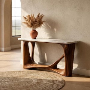 May include: A modern console table with a white marble top and sculptural, dark wood base. A terracotta vase with dried pampas grass sits on the table. A round, woven jute rug is on the floor.