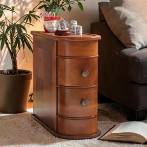 May include: A wooden side table with three drawers and ornate metal knobs. The table is a warm brown color and has a curved top and base. A snow globe and other decorative items sit on top of the table. A potted plant is to the left.