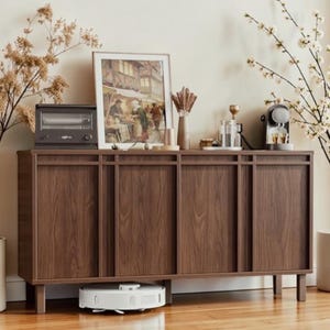 May include: A dark brown wooden cabinet with four doors, a framed picture, and a small toaster oven on top. A white robot vacuum cleaner is under the cabinet. Decorative plants and flowers are on either side of the cabinet.