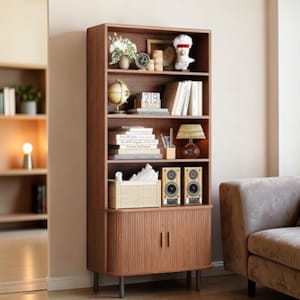 May include: A tall, brown wooden bookcase with multiple shelves. The shelves display books, decorative items, and a small lamp. The bottom section has a cabinet with two doors. The bookcase is set against a neutral wall, with a glimpse of a sofa.