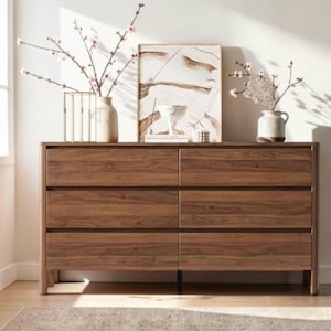May include: A rectangular wooden dresser with six drawers arranged in two columns of three. The dresser features a rich brown wood grain finish and rounded edges. On top sits a framed abstract painting, two ceramic vases with flowering branches.