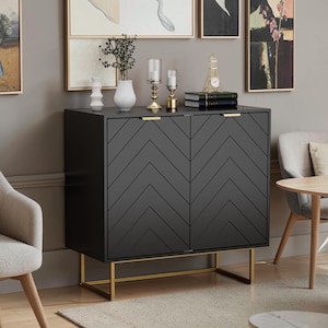 May include: A black rectangular cabinet featuring two doors with a chevron pattern and gold-toned metal handles. The cabinet is supported by a gold-toned metal frame base. The top surface holds a white vase, a plant, two candle holders, and books.