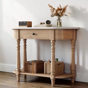 May include: A light-colored wooden console table with a drawer and a lower shelf. The table has turned legs and a white top. Two woven baskets sit on the lower shelf. A vase of dried flowers and a small clock are on the top.