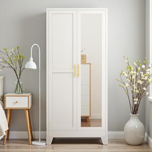 May include: A tall white wardrobe featuring two doors, one of which has a full-length rectangular mirror. The wardrobe includes two vertical gold-toned handles and is supported by four short, sturdy legs at the base, providing a clean, functional storage unit.