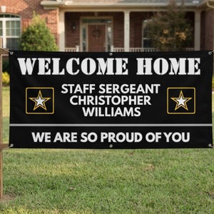 Op de afbeelding: Een zwarte rechthoekige banner met witte tekst die WELCOME HOME, STAFF SERGEANT CHRISTOPHER WILLIAMS en WE ARE SO PROUD OF YOU leest. Het ontwerp bevat twee geel omrande ster-emblemen en metalen ringen voor bevestiging aan een houten frame.