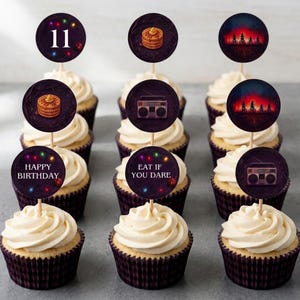 May include: A selection of cupcakes with white frosting and dark purple cases. Each cupcake is decorated with a circular image on a wooden stick. The images include the number 11, pancakes, a boombox, and the text "Happy Birthday" and "Eat if you dare."